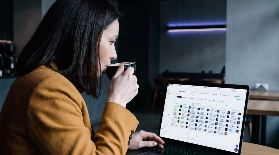 A woman in a moody cafe looks at a weekly content calendar on her laptop screen.